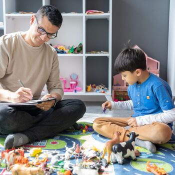 Little boy playing with toys with therapist with a clipboard sitting on the floor